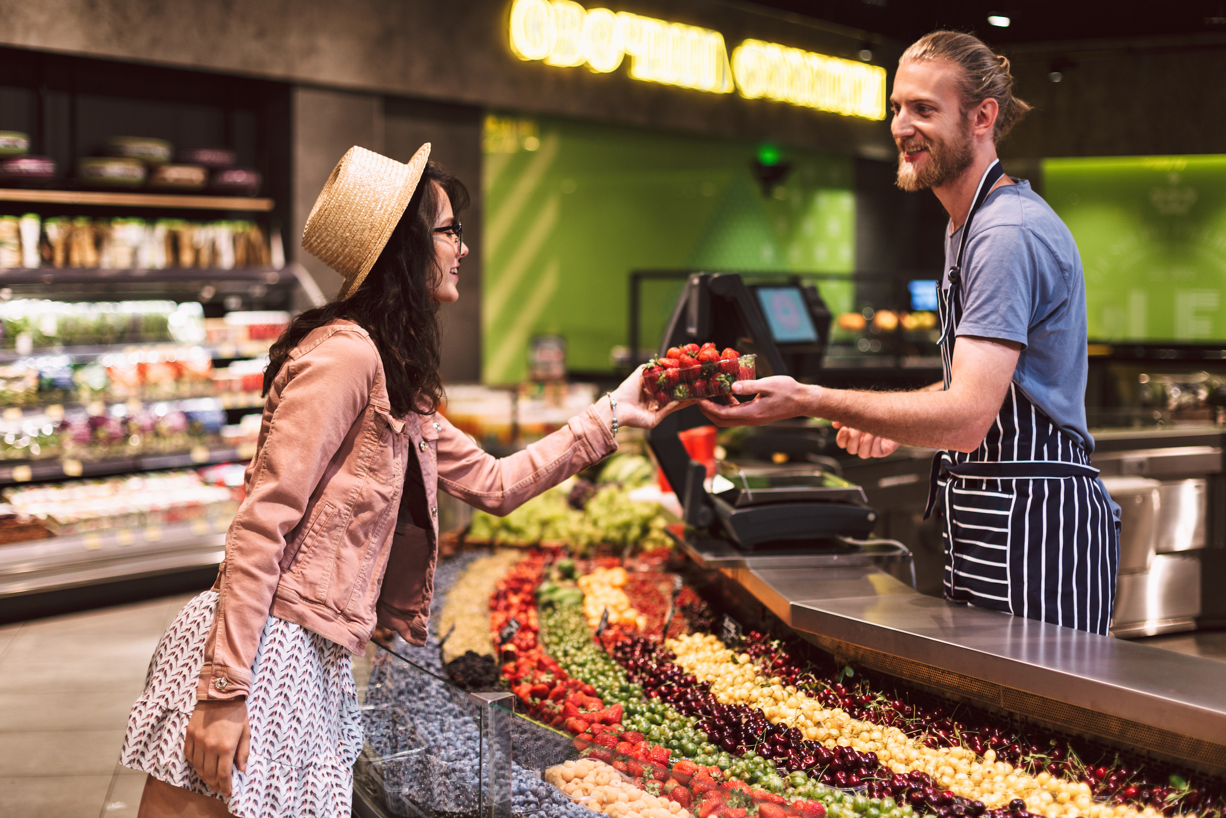 Young smiling seller in apron behind counter happily giving strawberries to customer in supermarket